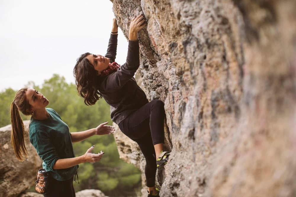 Two women rock climbing, with one behind the other with her arms raised up in. order to catch the other woman who is ascending, in piecer about customer trust.