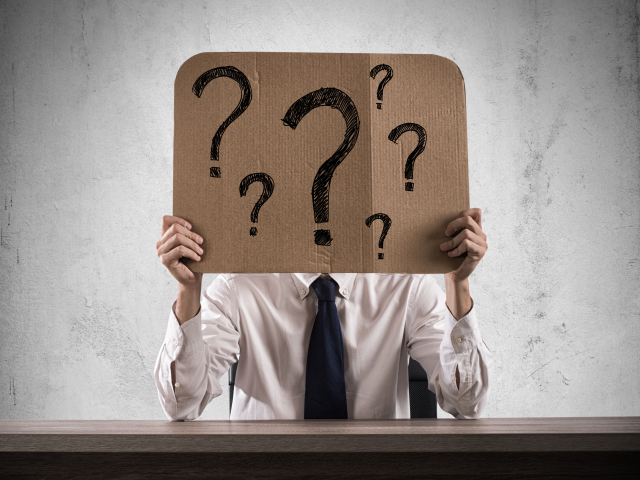 A businessman in the office holds a piece of cardboard with question marks over his face