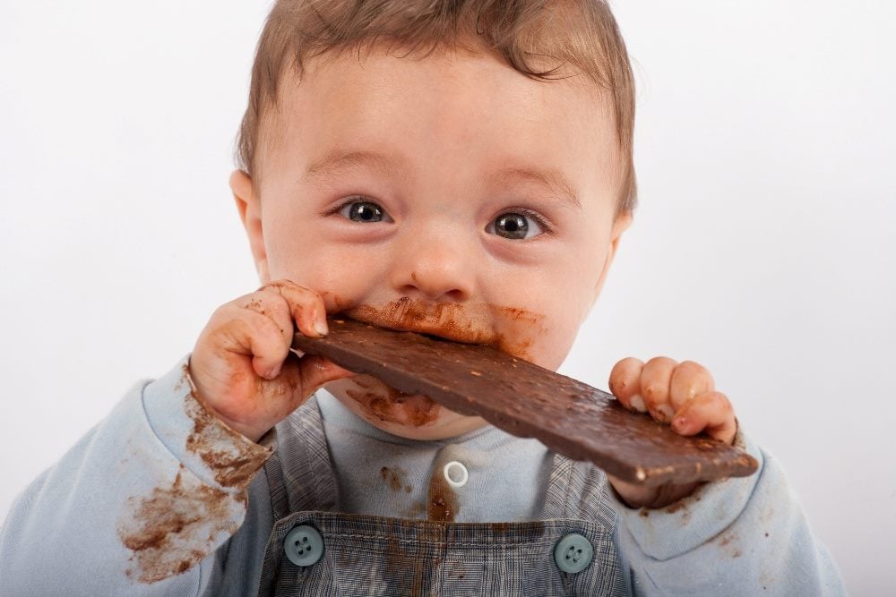 Cute baby wearing bib overalls is eating chocolate and has a messy face and hands in piece about customer experience strategies and peak experiences.