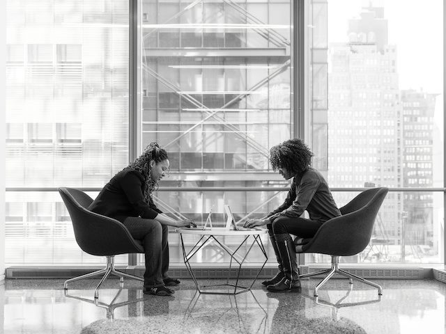 two women working facing each other, in front of window with view of high rise behind