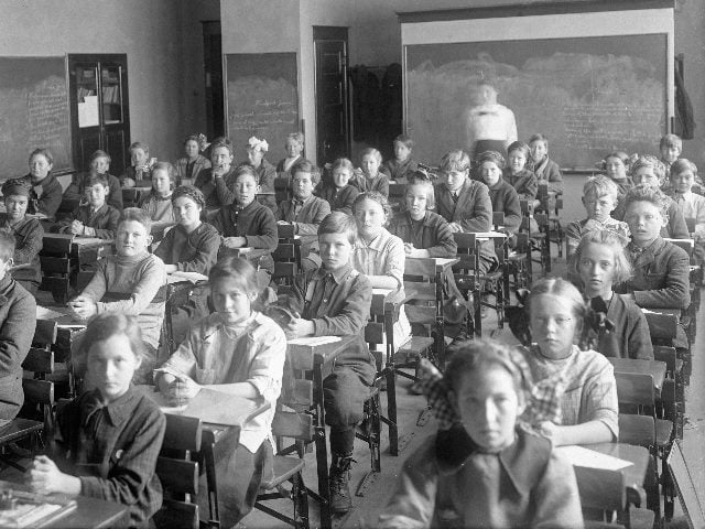Children sitting in an old-fashioned classroom in black-and-white photo.