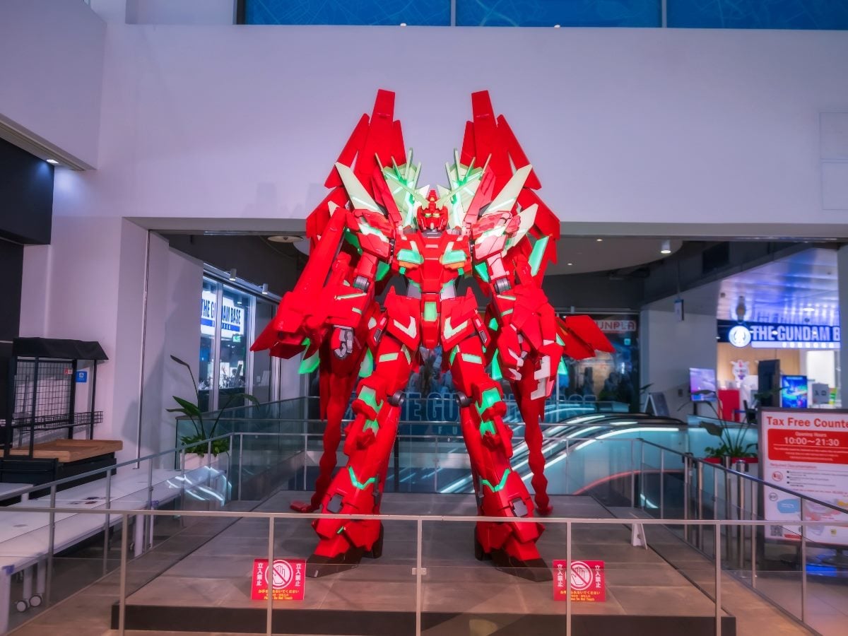 A large, brightly lit red and green Gundam-style robot statue displayed inside a shopping mall or entertainment complex. The life-size mecha figure stands on a platform surrounded by railings, with shops and signage visible in the background.