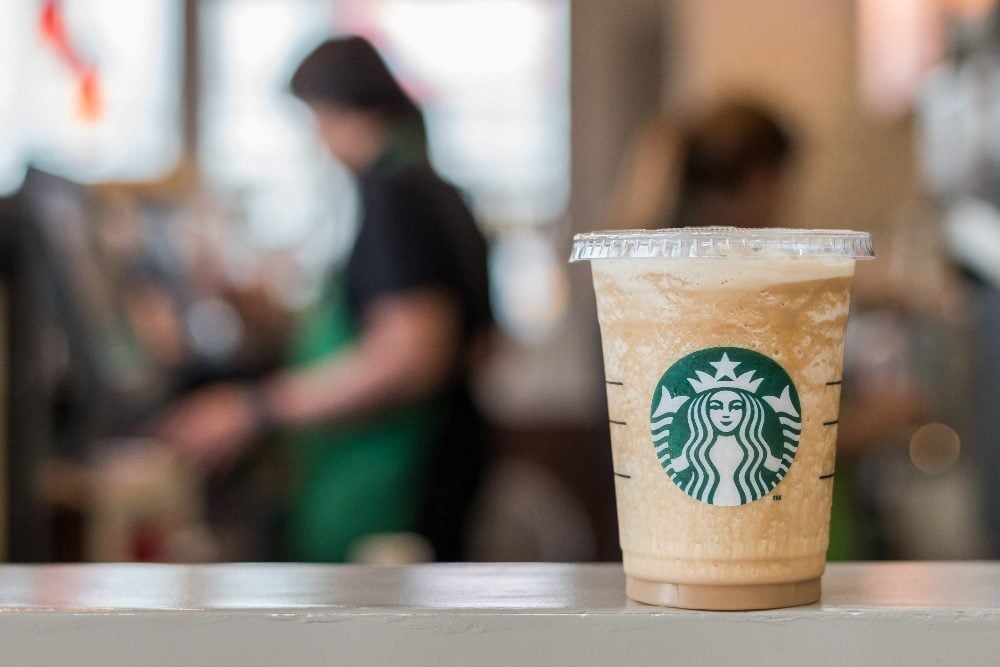 A Starbucks drink in a cups displaying the green and white logo sits on a table at a Starbucks store with a barista working in the blurred background in piece about Starbucks protests and PR crisis management.