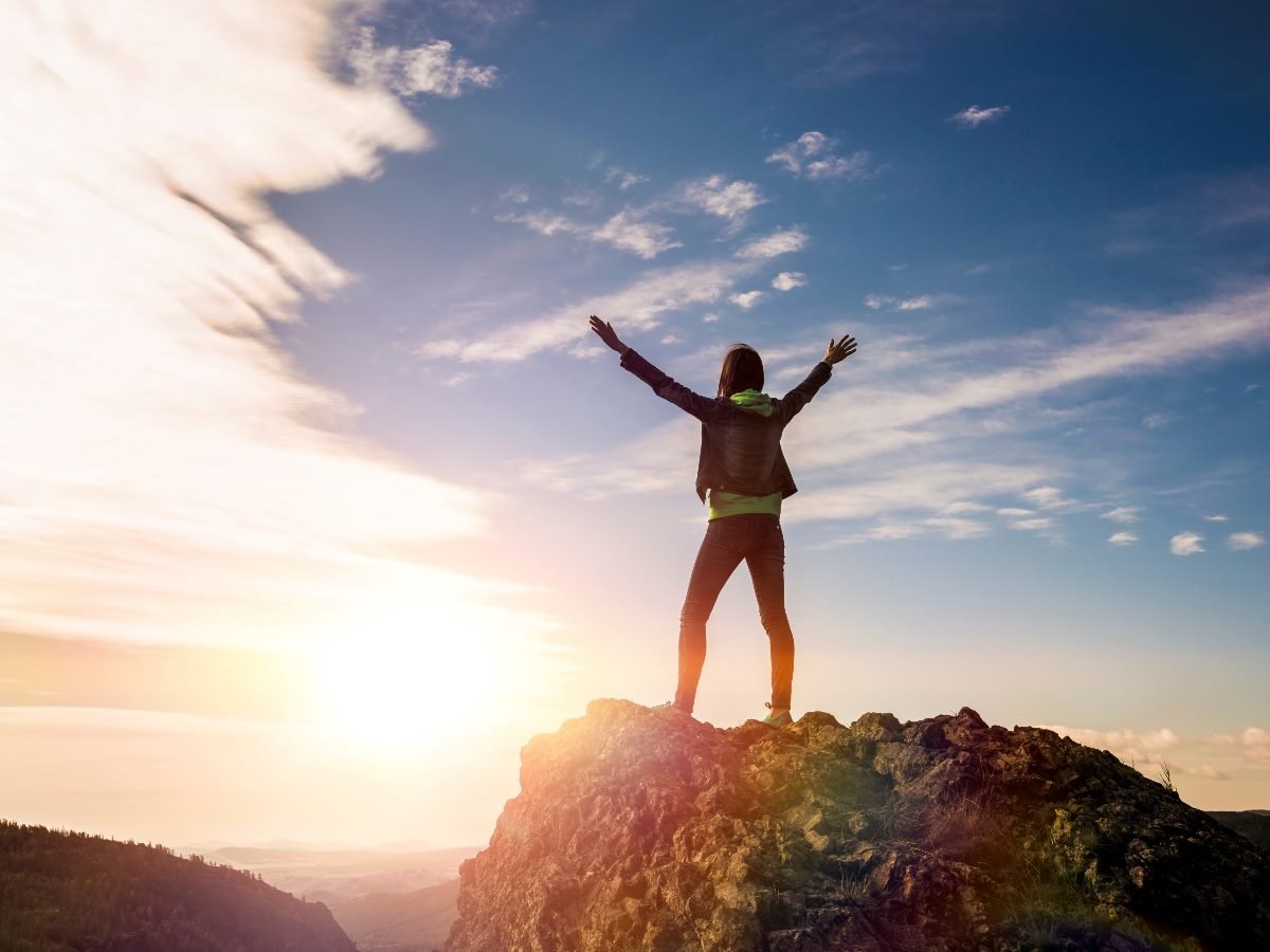 Woman stand on top of a mountain with arms raised in celebration.