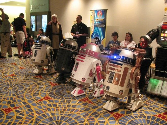 A row of four R2D2s robot models from the movie Star Wars in a movie theater lobby surrounded by people.