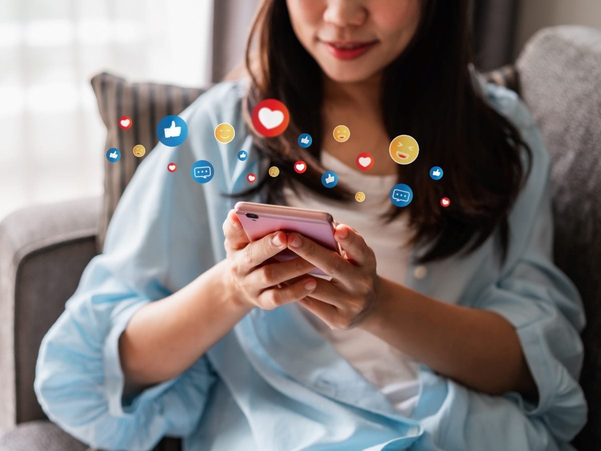 Person sitting on a couch using a smartphone while colorful social media reaction icons (likes, hearts, comments, emojis) float around the screen, illustrating digital engagement and interaction.