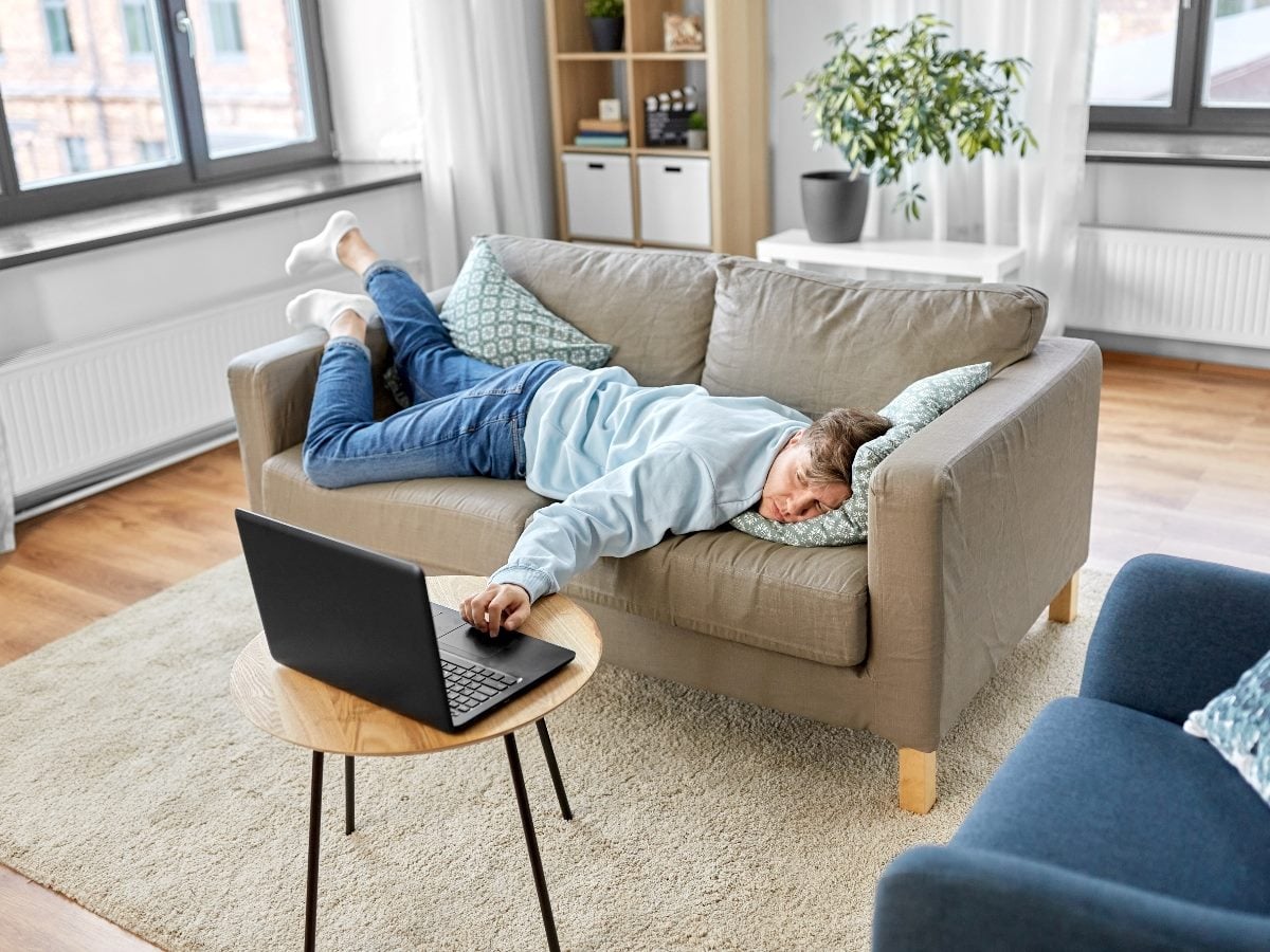 Person lying face-down on a couch in a bright living room, appearing bored or exhausted while lazily reaching toward a laptop on a small table.