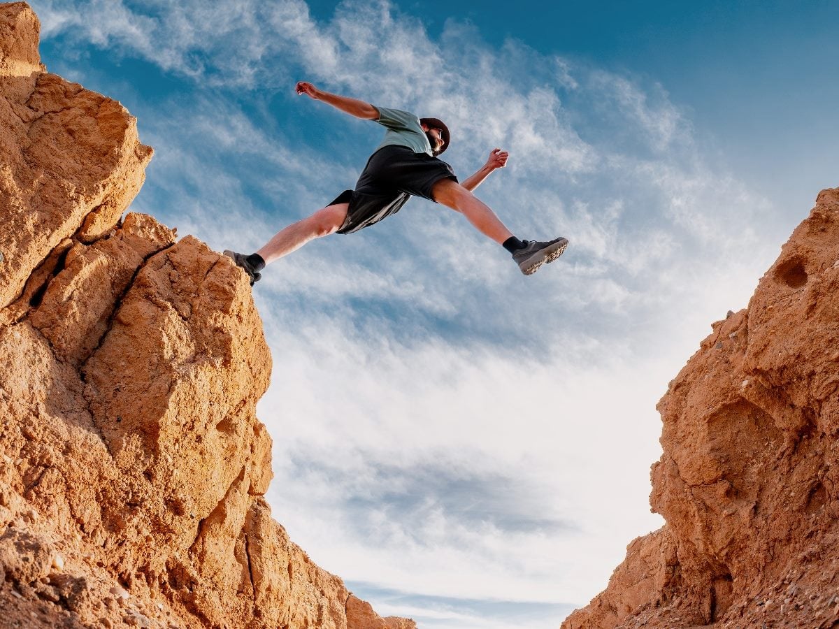 Low-angle view of a person mid-jump leaping across a wide rocky canyon, legs outstretched between two cliffs under a blue sky with scattered clouds, capturing a moment of risk and forward momentum.