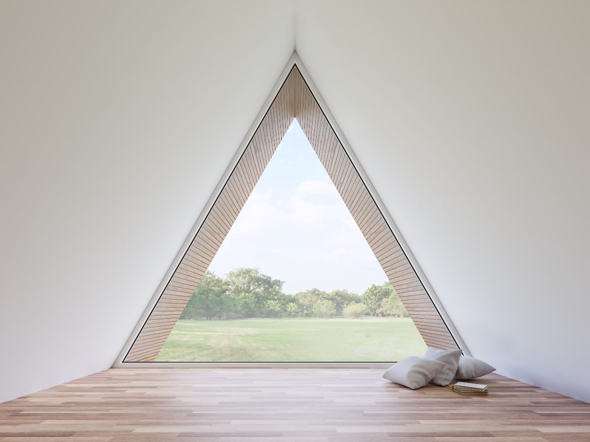 A minimalist interior with a large triangular window overlooking a grassy field and trees, with two pillows and a book placed on the wooden floor.