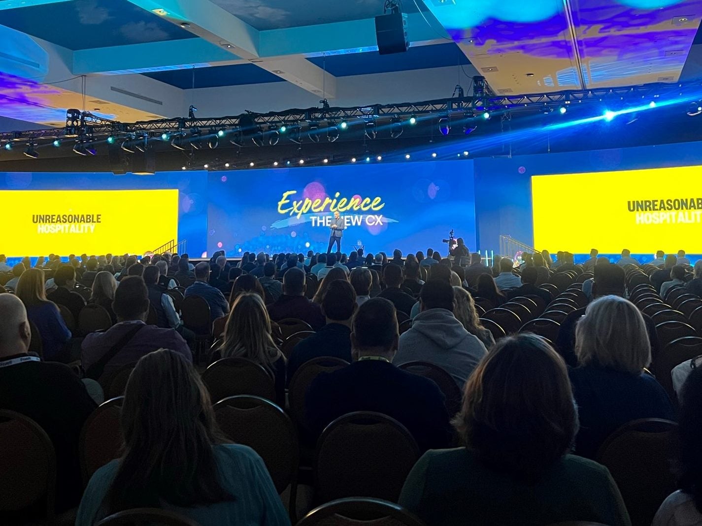 “Audience fills a large ballroom as a keynote speaker presents on the main stage at the Five9 customer conference, with screens reading ‘Experience the New CX’ and ‘Unreasonable Hospitality.’”