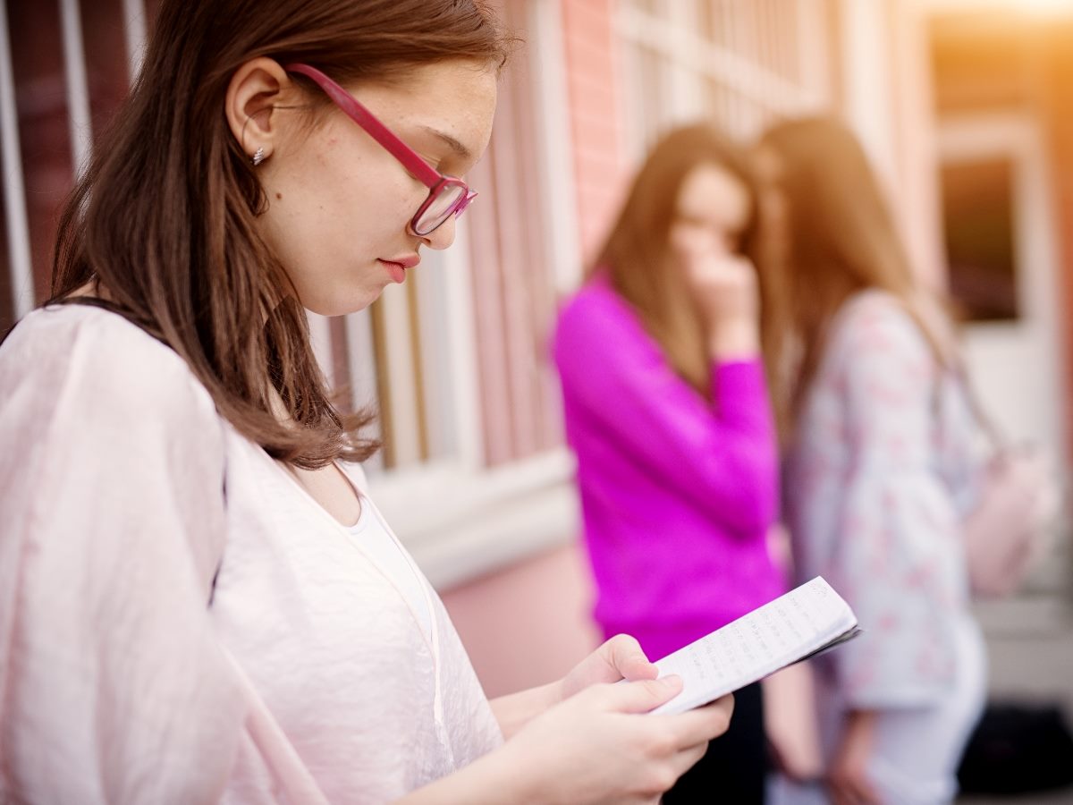 Sad high school girl reading from school notes in foreground while her schoolmates gossip her in background while standing outside of the school. 