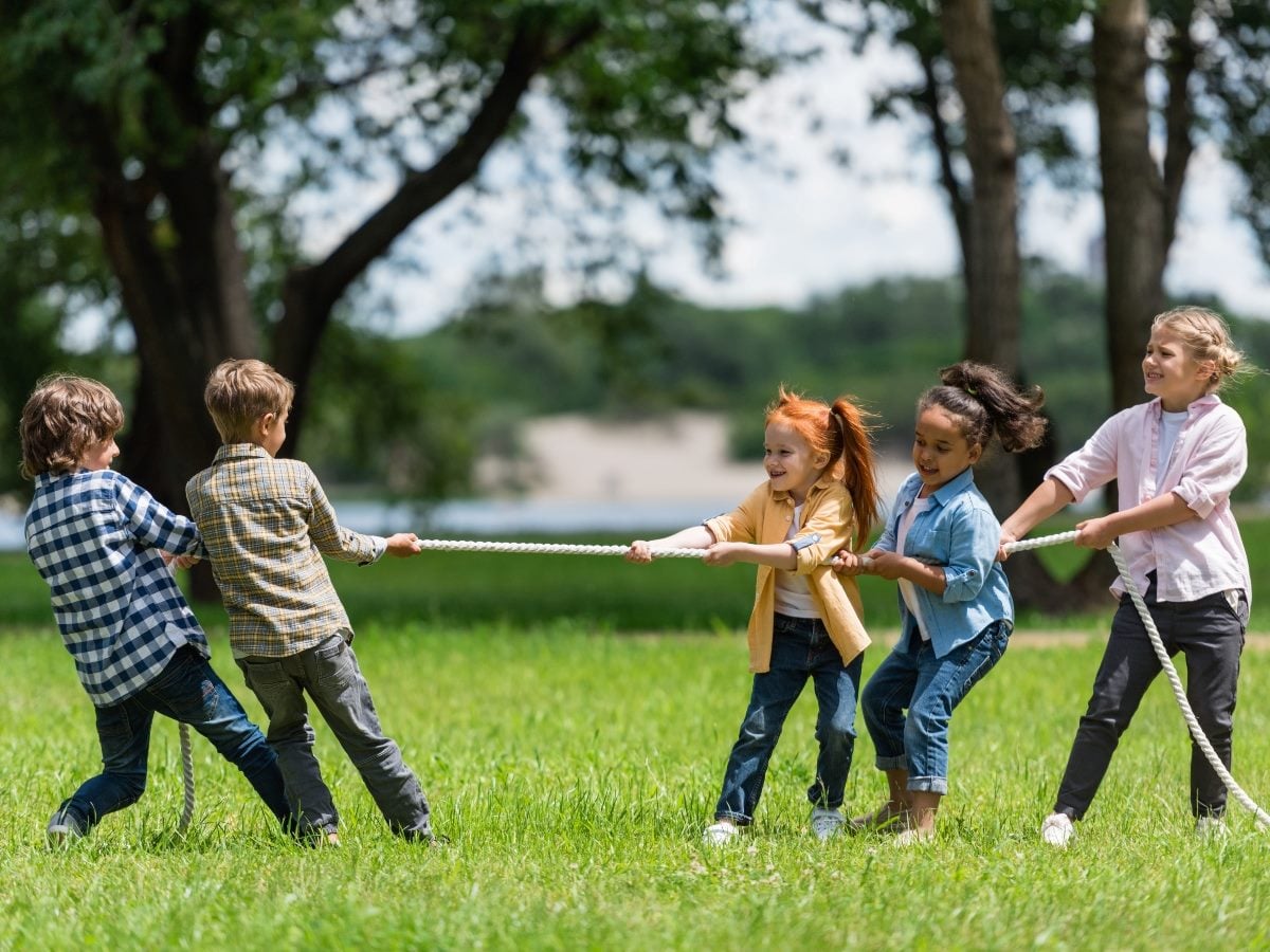 Five children play tug-of-war on a grassy field in a park, smiling and pulling on opposite ends of a rope. Large trees frame the scene, with water and a wooded shoreline visible in the background on a sunny day.