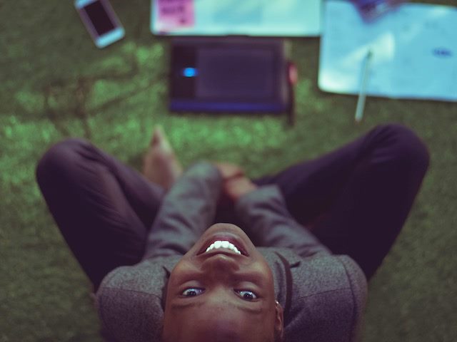 woman seated on the floor with multiple devices and work in front of her, looking up at the  camera