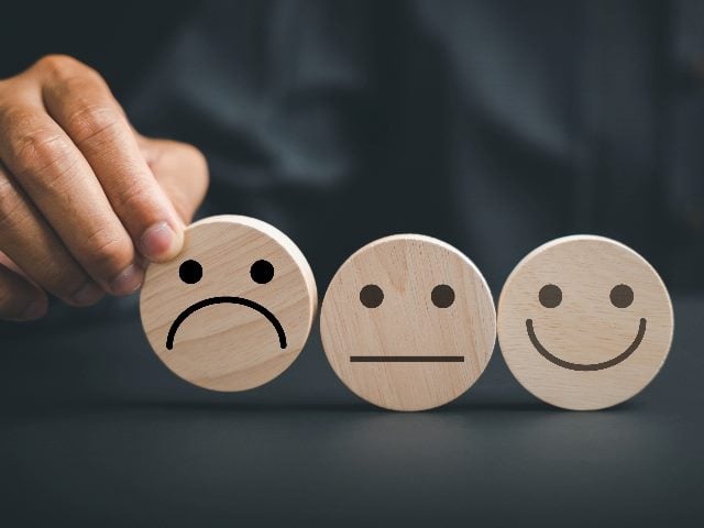 Negative customer experience concept. A hand picking up a frown icon on wooden circle, next to two other wooden circles with reactions: expressionless and happy.