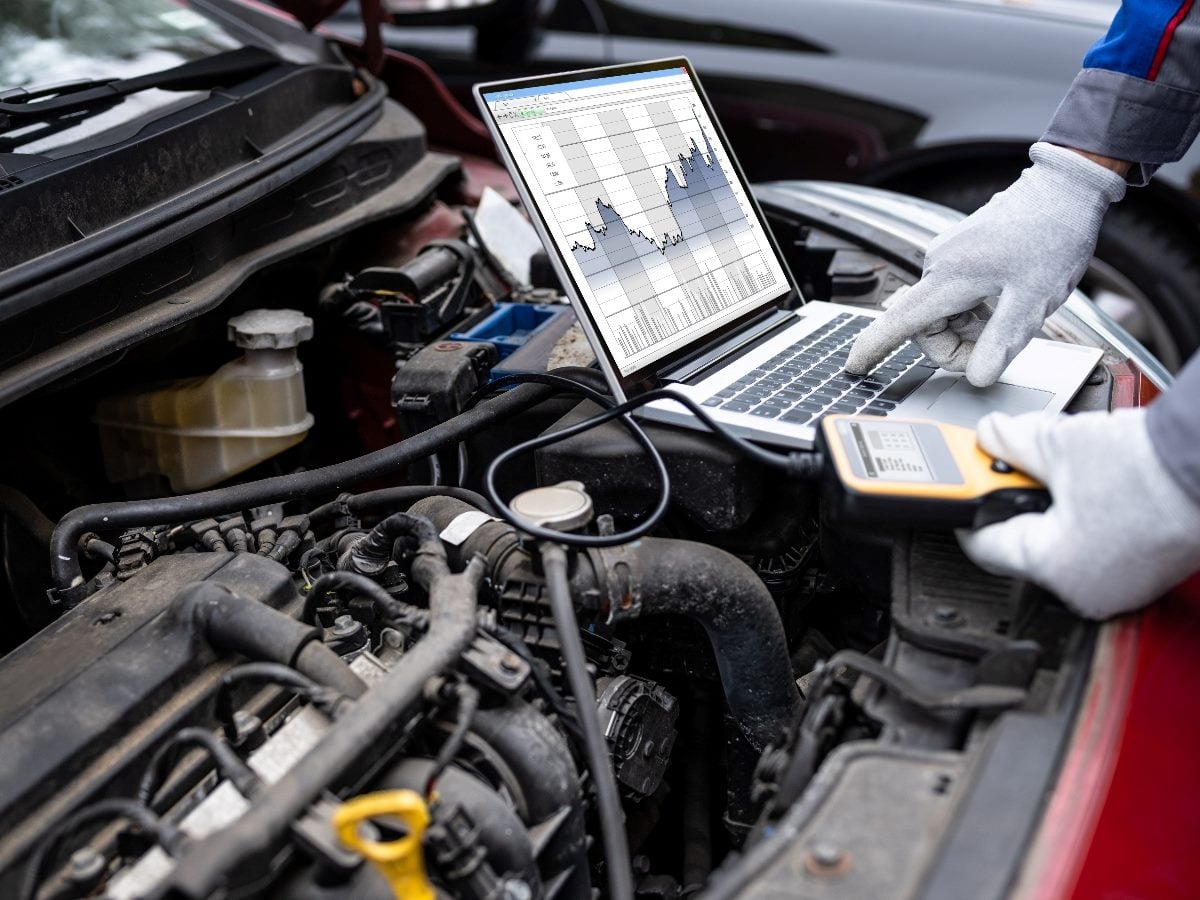 Technician’s gloved hands using a laptop to run diagnostic analysis on a car engine, reviewing performance data while connected to testing equipment.