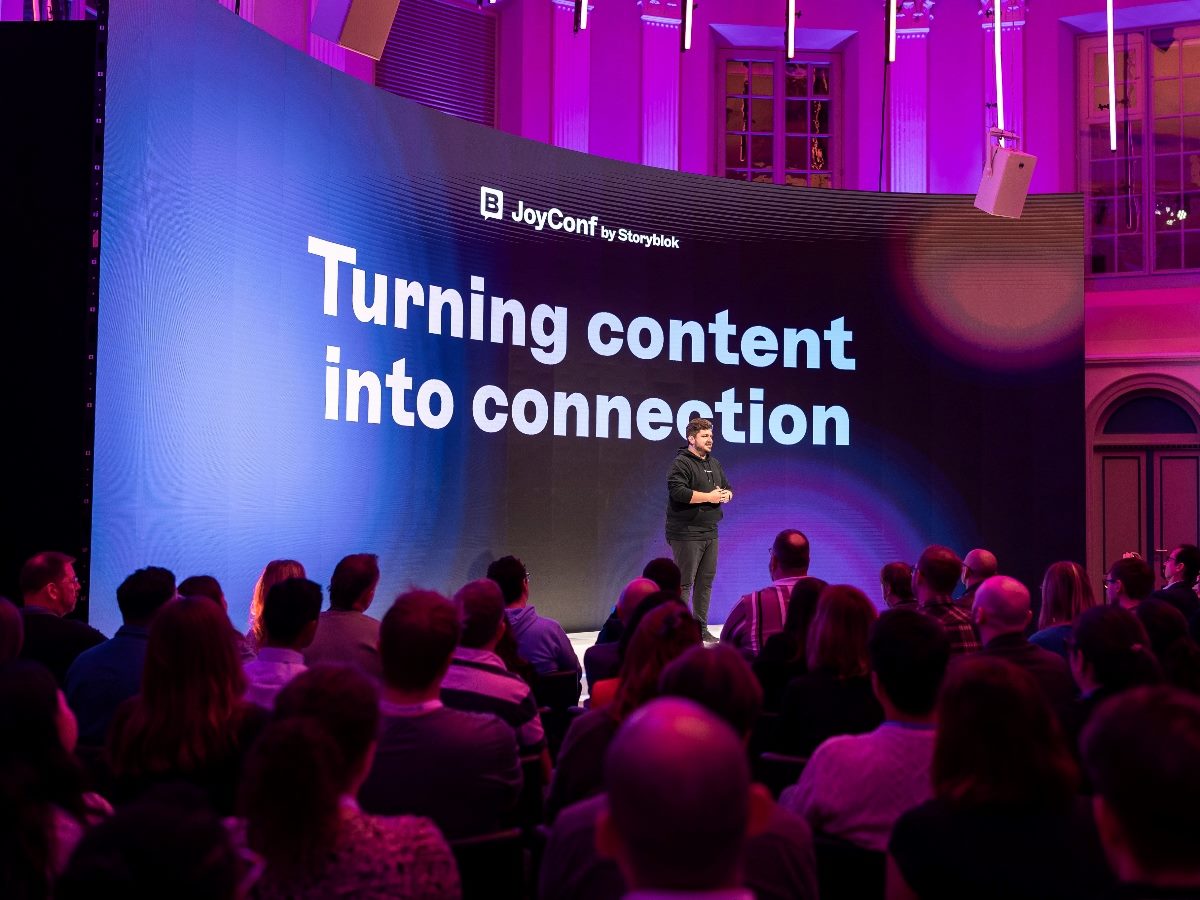 Dominik Angerer, CEO of Storyblok, stands on stage at JoyConf in front of a large screen displaying the words “Turning content into connection,” speaking to an audience in a brightly lit venue with purple and pink lighting.