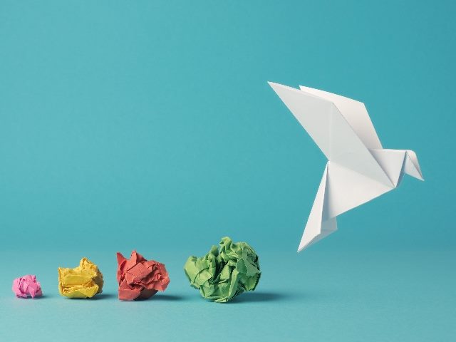 A pink, yellow, red and green balls of wadded up papers sit on a blue surface and background an a white paper bird arises, suggesting the process of business transformation.