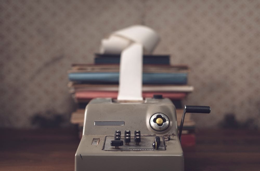 A vintage calculator with a paper roll sits on a desk, backed by a stack of old books. This image represents the traditional methods of calculating and demonstrating marketing ROI, emphasizing the need for updated, integrated approaches in modern marketing.