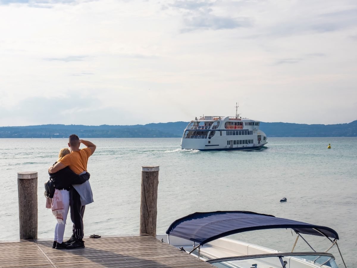 A couple stands arm in arm at the edge of a wooden dock, watching a white passenger ferry glide across a calm bay on a hazy day, with a small motorboat moored in the foreground.