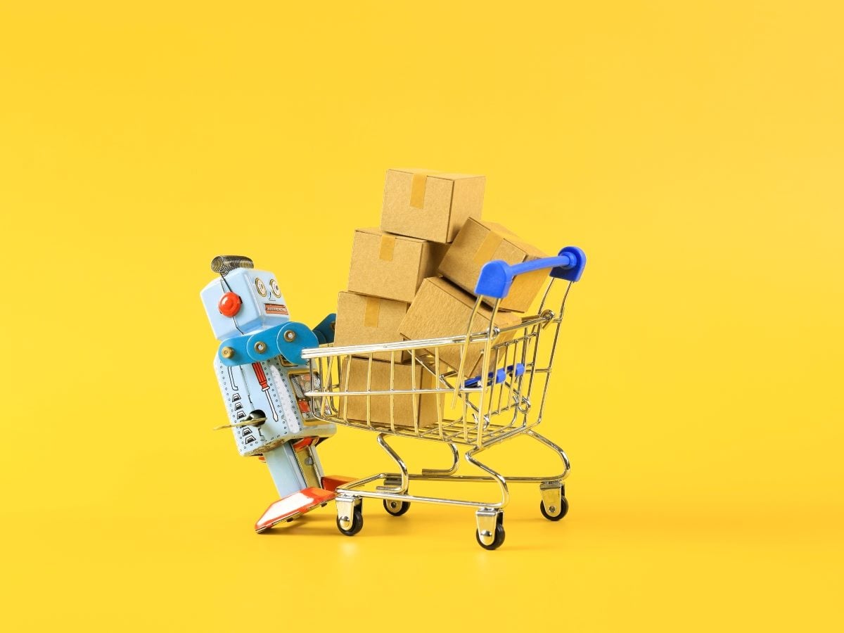 A toy robot leans against a miniature shopping cart filled with stacked cardboard shipping boxes on a bright yellow background, symbolizing AI, automation and ecommerce fulfillment.