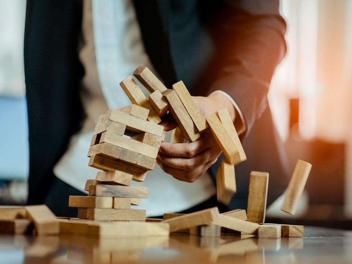 Business professional in a suit holding collapsing wooden blocks from a Jenga-style tower as pieces fall onto a table, symbolizing instability or organizational breakdown.