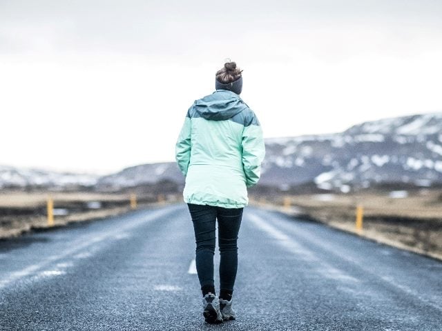 Shot from behind a woman walking on an empty road with the backdrop of snowy mountains.