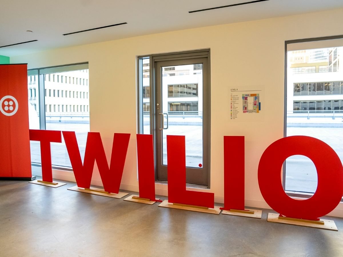 Large red Twilio logo letters displayed inside a modern office space with floor-to-ceiling windows and a branded banner in the background.