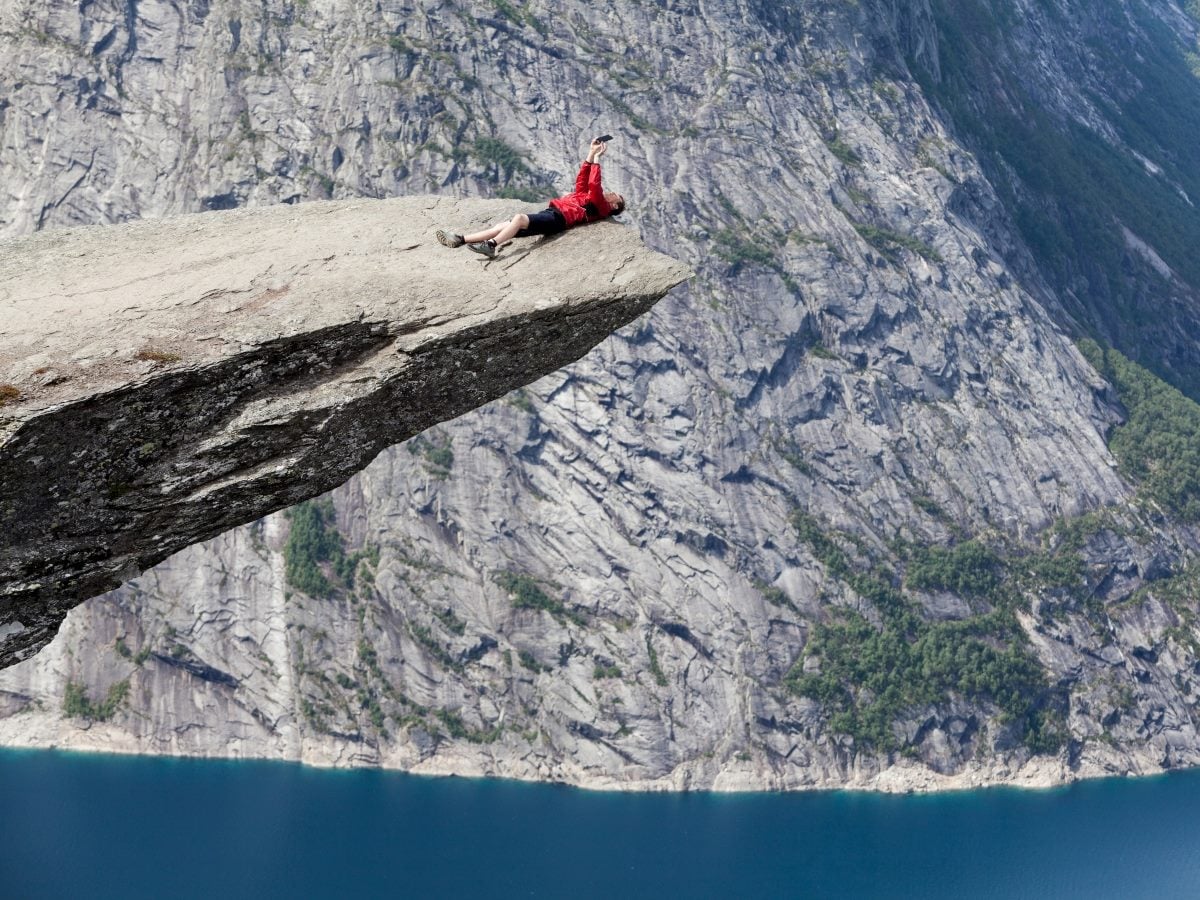 A person in a red jacket and black shorts lies on the edge of a dramatic rock outcrop high above a deep blue fjord, holding a smartphone overhead. Steep granite cliffs and patches of green forest fill the background, emphasizing the height and expansive mountain landscape.