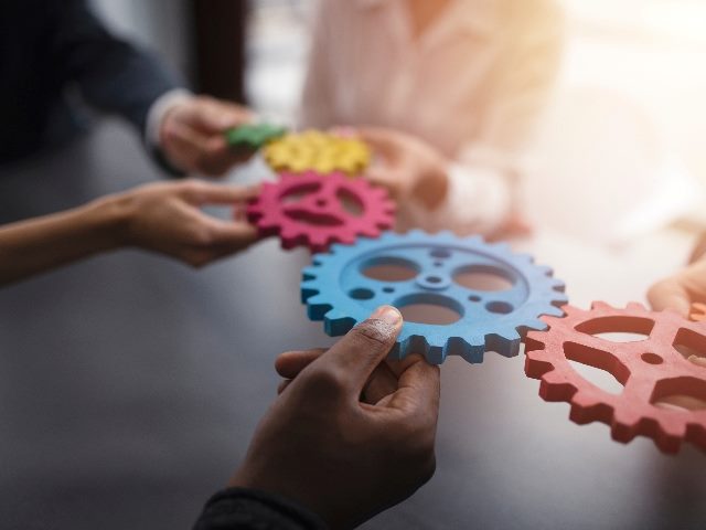 Hands of business team members hold multi-colored gears of red, yellow, green, blue and orange in piece about AI workplace integration.