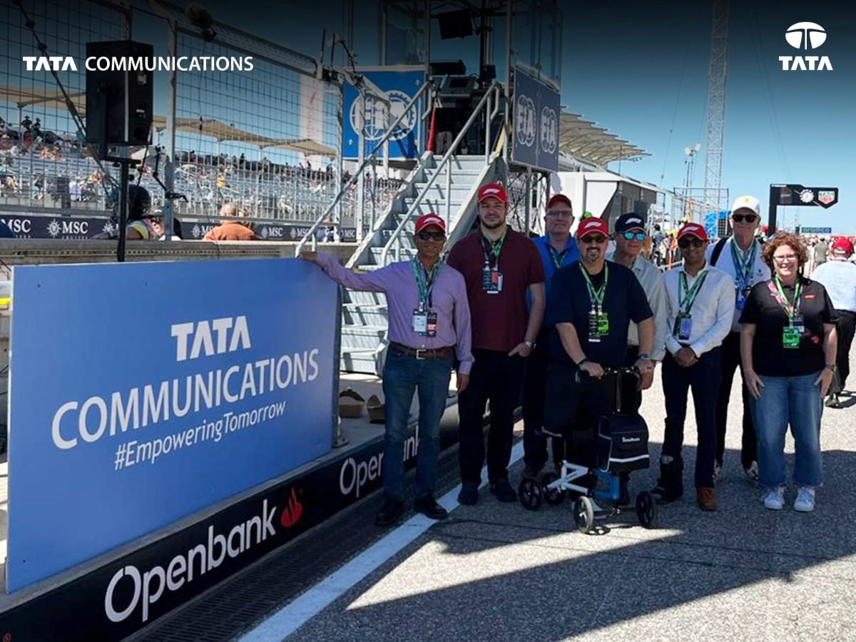 A group of people wearing event badges and red hats pose on a racetrack beside a large Tata Communications banner that reads “#EmpoweringTomorrow,” with grandstands and spectators visible in the background.
