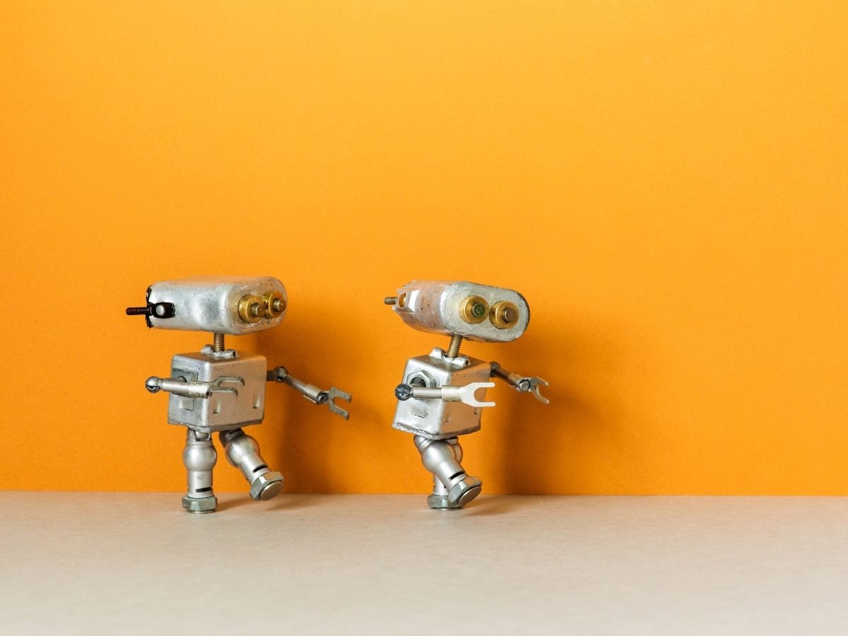 Two small silver toy robots made from metal parts stand side by side on a light-colored tabletop against a bright orange background, appearing to walk or interact with one another.