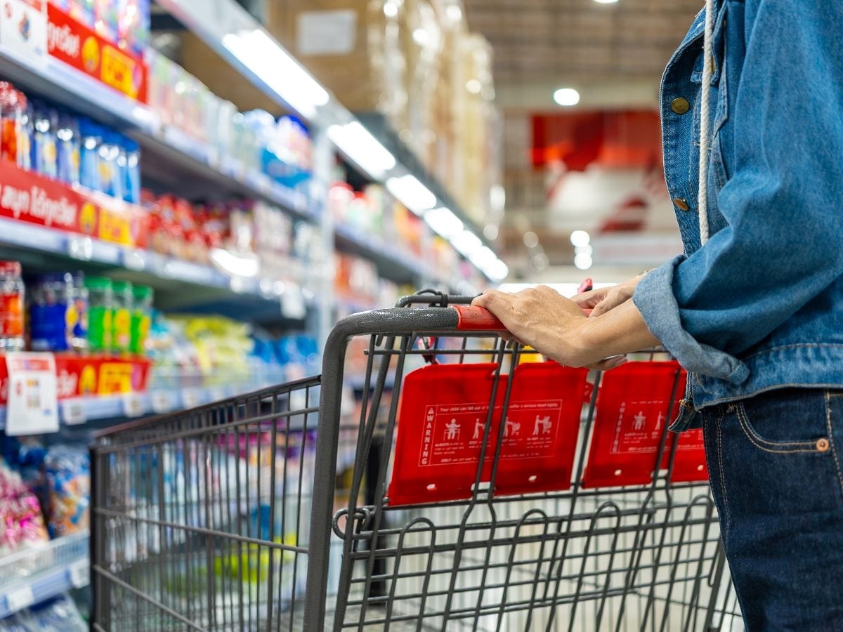 Person pushing a shopping cart down a brightly lit grocery store aisle. Shelves stocked with beverages, snacks and packaged goods line both sides of the aisle, while the shopper, dressed in a denim jacket and jeans, is shown from the side with hands on the cart handle. The background is softly blurred, emphasizing the retail shopping experience and consumer purchasing behavior.