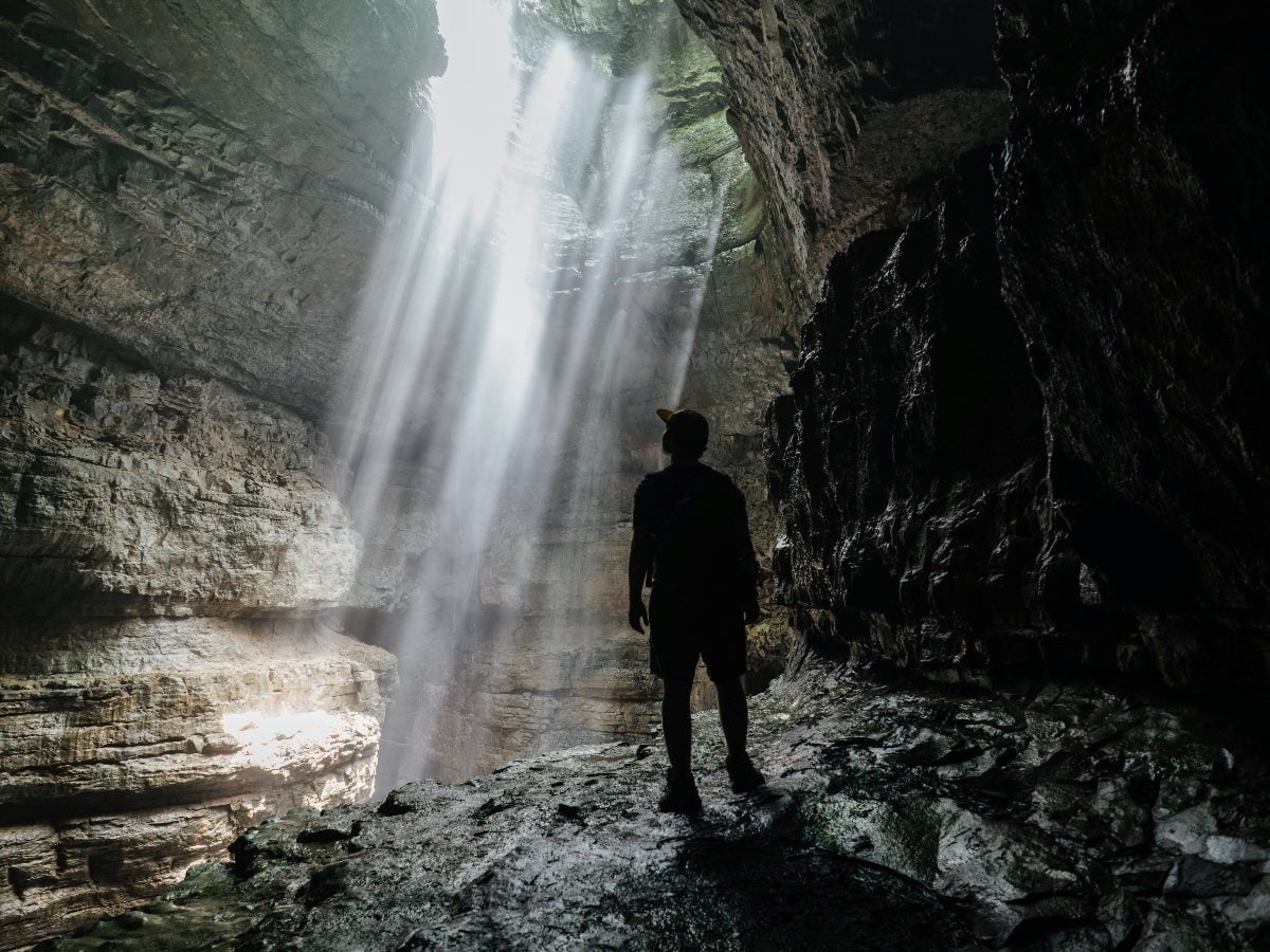 A silhouetted explorer stands inside a large cave as bright beams of sunlight stream through an opening above, illuminating wet rock walls and the cavern floor.