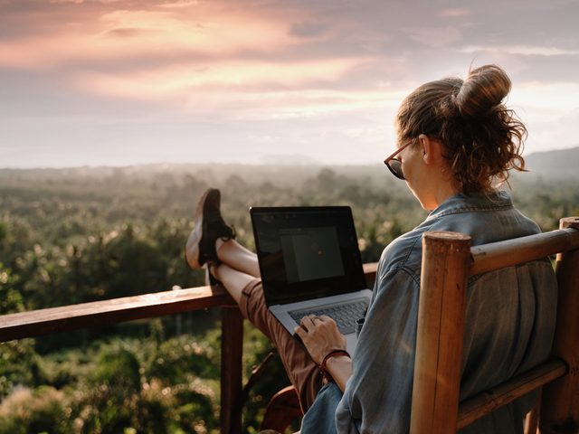 A woman sitting in a chair feet up on a deck railing working on a laptop.