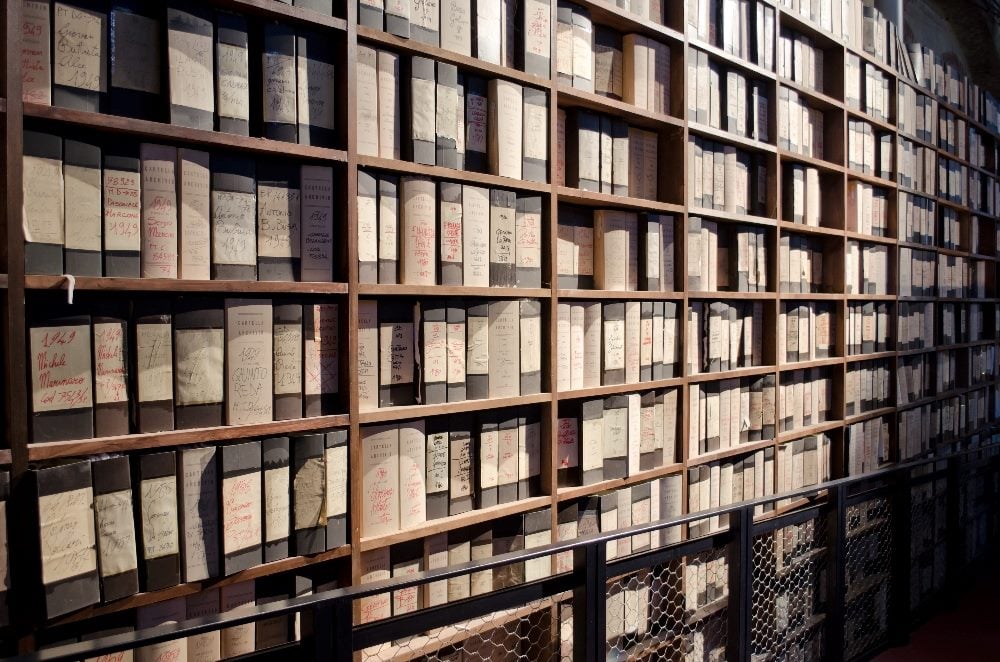 A library archive with numerous shelves filled with organized, labeled boxes and binders, showcasing a system of meticulous record-keeping and storage. The labels display handwritten notes and dates, indicating a historical collection in piece about the evolution of AI in DAM (digital asset management).