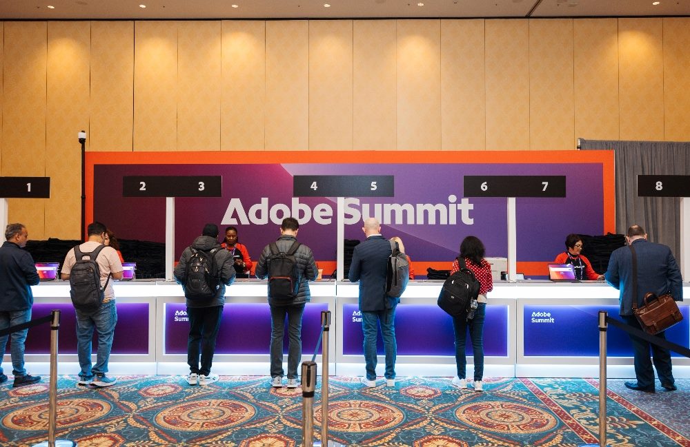 Seven people line up to get registered at the Adobe Summit 2024 registration desk that says, "Adobe Summit" on a sign.