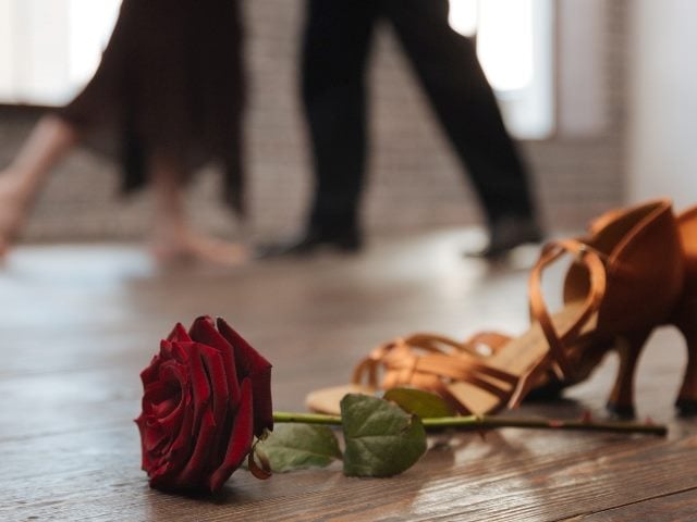 The image captures a moment on a wooden dance floor. In the foreground, a vivid red rose lies beside a pair of abandoned tan dance shoes. The background is softly blurred, showing the motion of two dancers' feet as they glide across the floor dancing the Tango in piece about Apple’s release of AI features at WWDC 2024.
