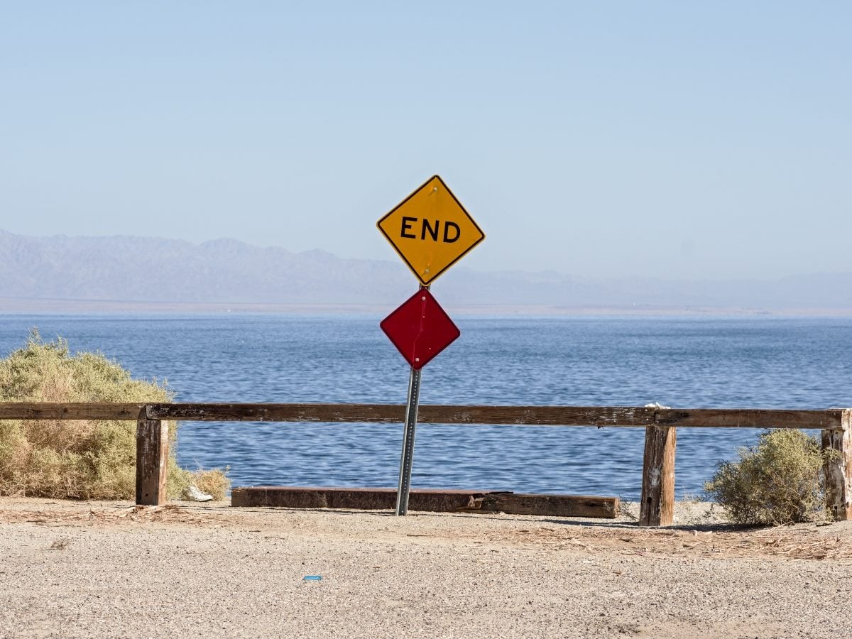 A yellow diamond road sign reading "END" sits atop a red diamond warning sign, mounted on a metal pole in front of a low wooden guardrail. Behind the sign, a calm blue lake stretches to a hazy line of distant mountains under a clear sky. Dry scrub brush grows on either side of the fence, and the foreground is bare gravel and sand.