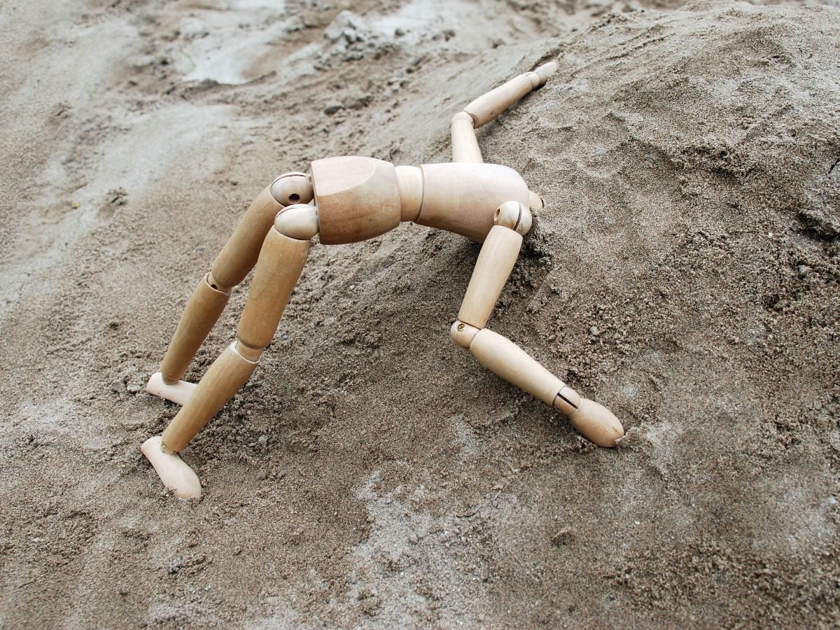 A wooden artist mannequin is sprawled face-down on a sandy mound, positioned as if it has collapsed or is struggling to climb the hill.