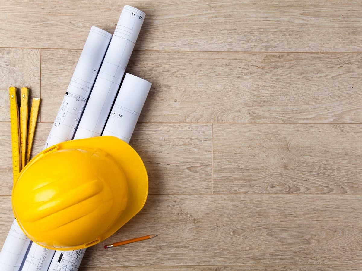 Yellow construction hard hat resting on rolled architectural blueprints and measuring tools on a light wood surface, symbolizing planning and structural design.