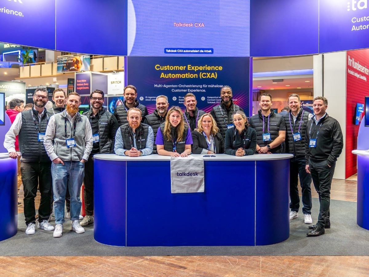 Talkdesk team members stand at a Customer Experience Automation (CXA) booth during a technology trade show, with large digital displays highlighting Talkdesk CXA and multi-agent orchestration for customer experience.