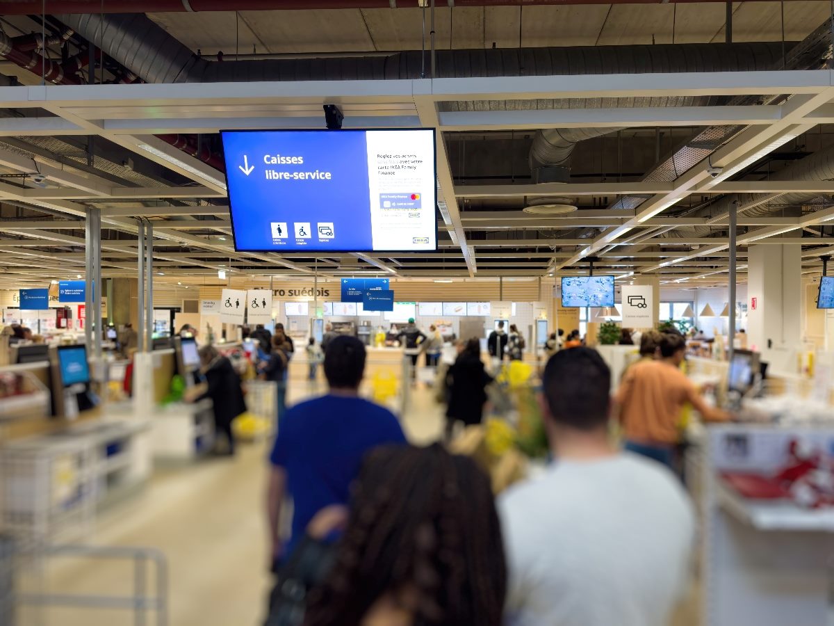 Customers standing in line at a self-service checkout area in a large retail store, with an overhead sign in French indicating “Caisses libre-service” (self-service checkouts).