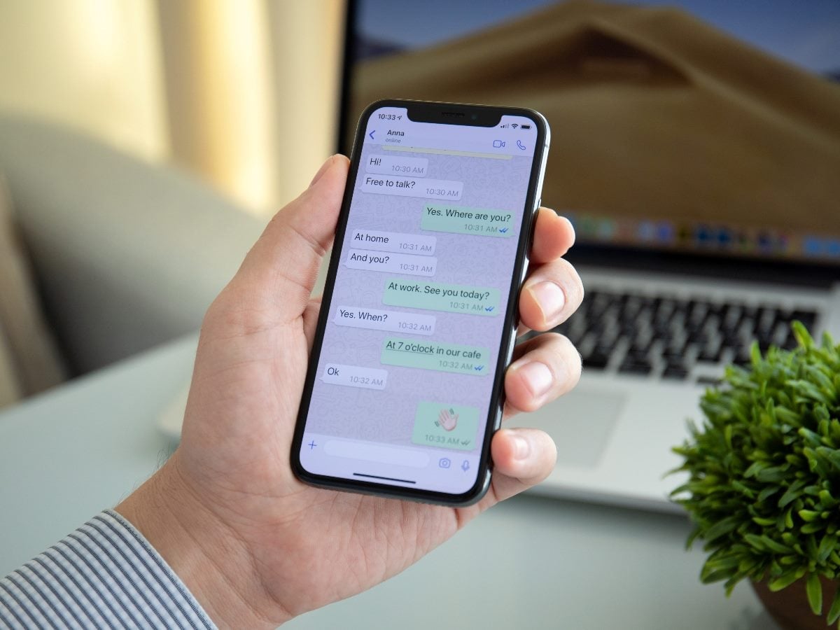 A person holds a smartphone displaying a WhatsApp-style text conversation, with a laptop and desk workspace blurred in the background, illustrating everyday messaging on a mobile device.
