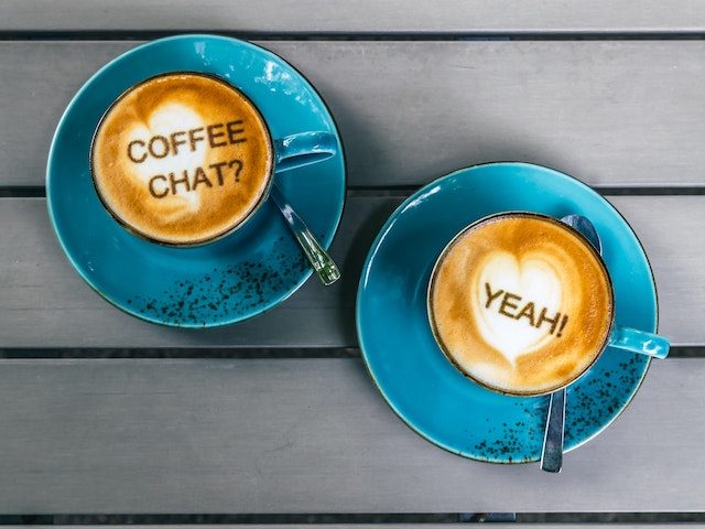 Two side-by-side turquoise coffee cups in their saucers with foam that reads "Coffee Chat?" and "Yeah!!"