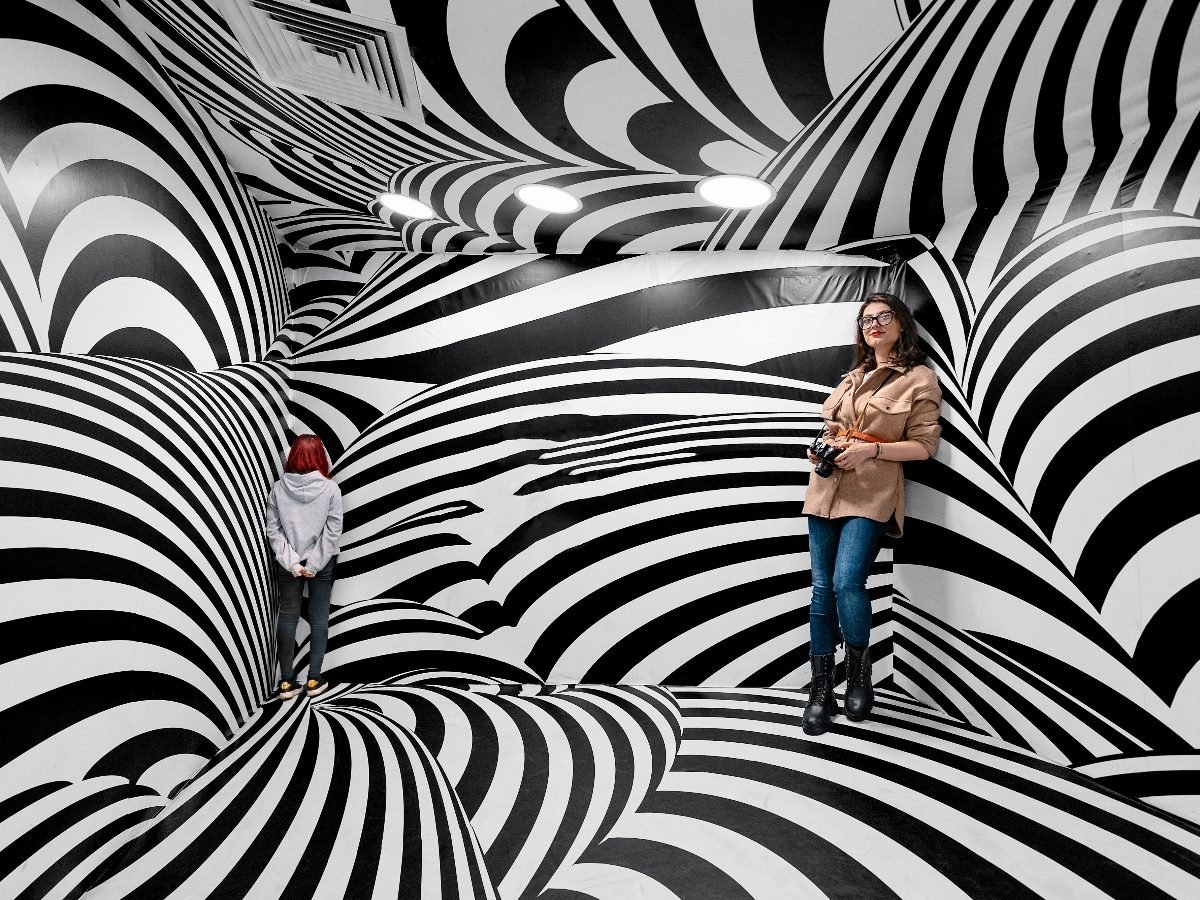Two people stand in a black-and-white, optical illusion room covered in swirling striped patterns that distort perspective; one faces the wall while the other leans against it holding a camera.