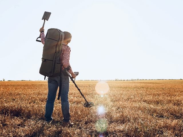  Man with metal detector equipment searching for metal goods in a field.