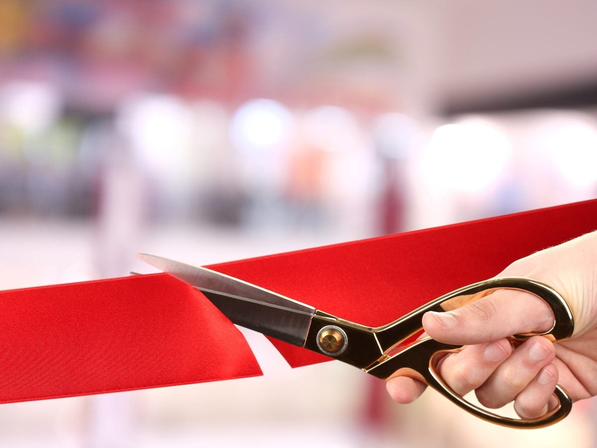 A close-up of a hand holding gold ceremonial scissors cutting a bright red ribbon, symbolizing an opening or launch event.