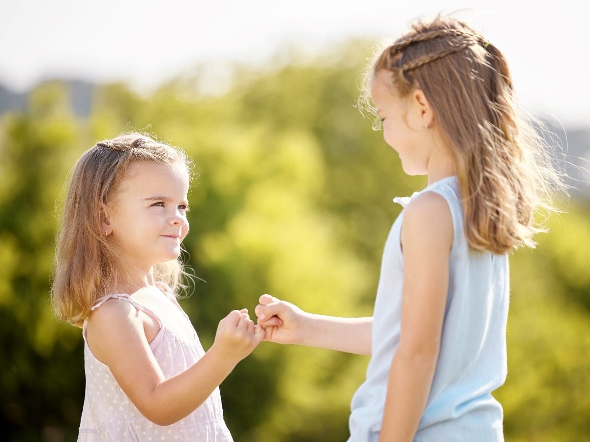 Two young girls standing outdoors in bright sunlight, smiling and linking pinky fingers while facing each other, with blurred green trees in the background.