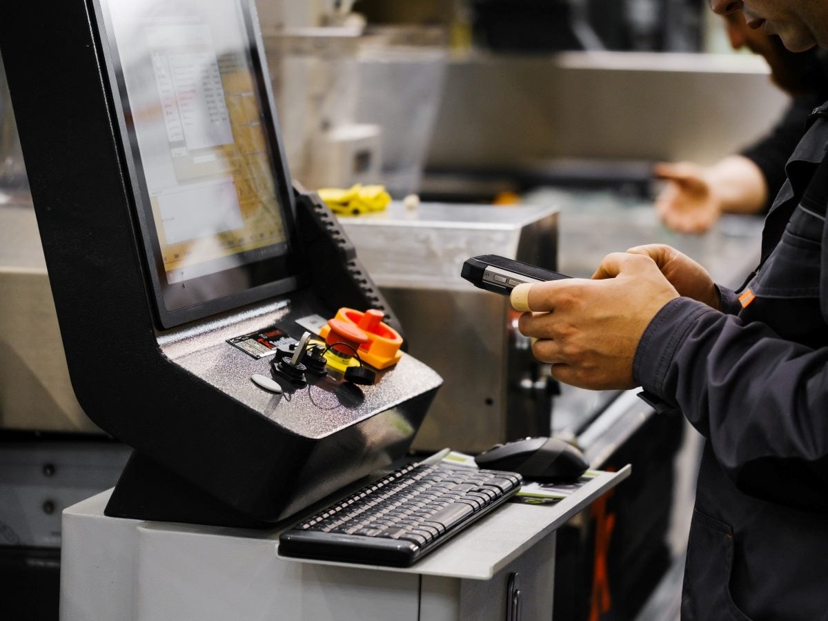 A worker in an industrial manufacturing environment uses a handheld barcode scanner while standing at a workstation with a computer terminal, keyboard and mouse. The image highlights digital operations, inventory tracking and shop floor data collection in a production setting.