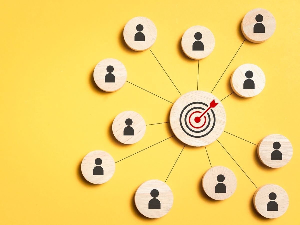 Wooden circular icons representing people arranged around a central wooden disc with a red bullseye target and arrow, connected by thin lines on a yellow background, symbolizing customer signals converging toward strategic focus.